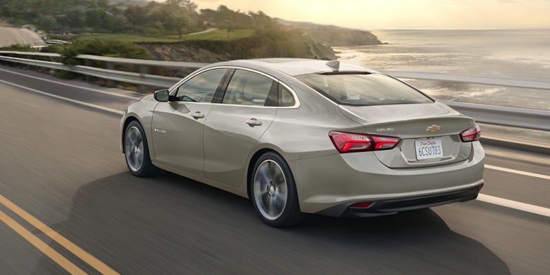 A Chevrolet Malibu driving along a scenic road surrounded by trees and blue sky.